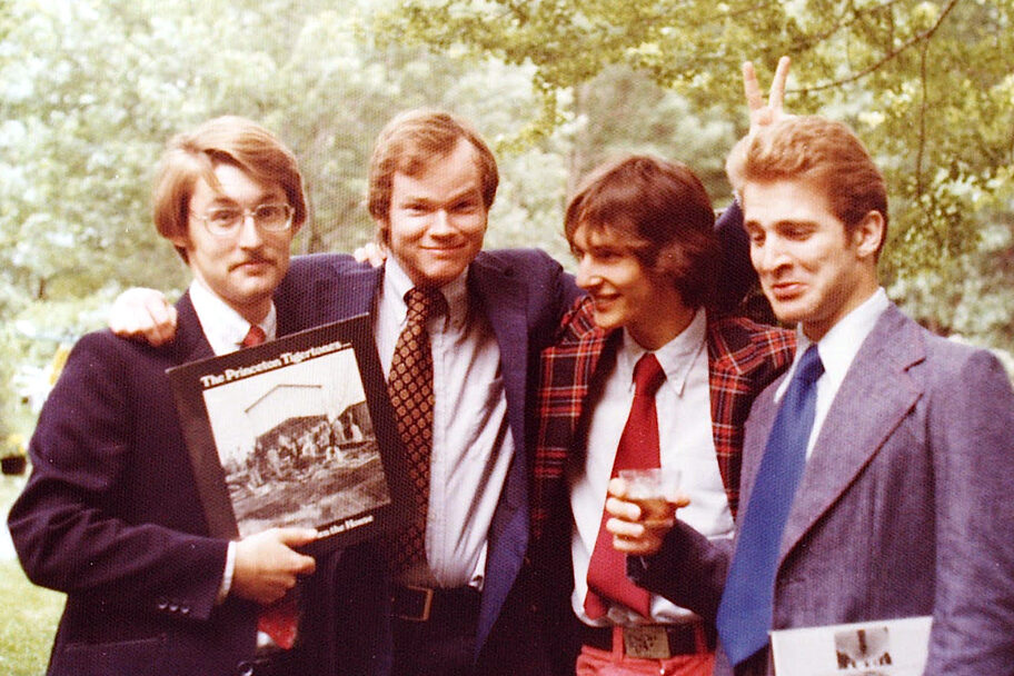 Four young men in ties and ugly blazers holding record albums and cocktails. One of them is throwing bunny ears behind the head of another.